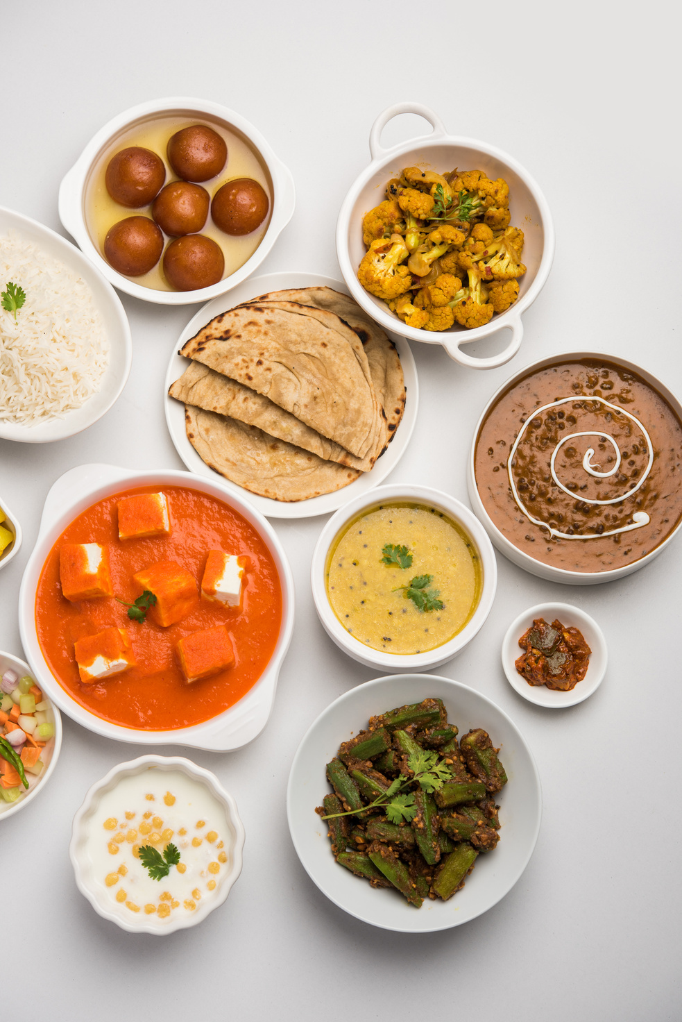 Assorted Indian food like paneer butter masala, dal, roti, rice, sabji, gulab jamun and bound raita served in bowls over moody background, selective focus